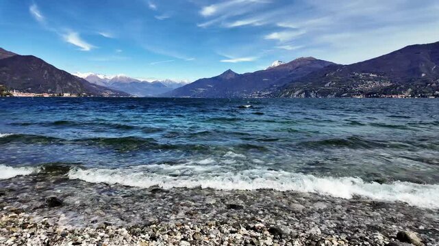 Panoramic view of lake como with waves on rocky shore and alpine mountains under blue sky