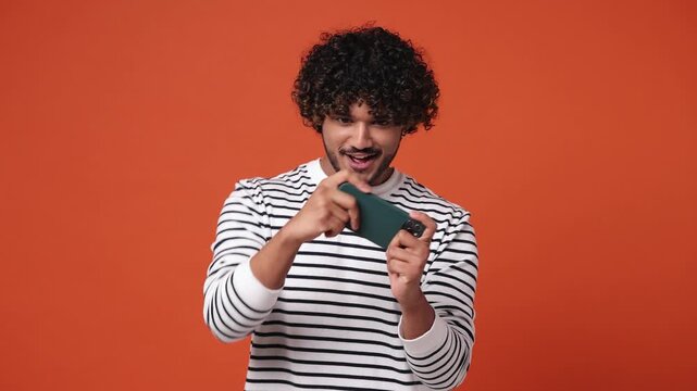 Gambling excitable young Indian man he wear striped sweatshirt casual clothes use play racing app on mobile cell phone hold gadget smartphone for pc video games isolated on plain orange red background