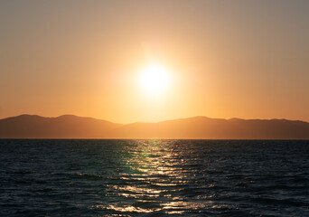 Naklejka premium Sun dips below distant mountains painting the vast ocean with a warm, fiery glow at dusk