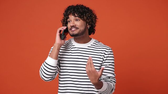 Young Indian man wear striped sweatshirt casual clothes hold use talk on mobile cell phone conducting pleasant conversation isolated on plain orange red background studio portrait. Lifestyle concept