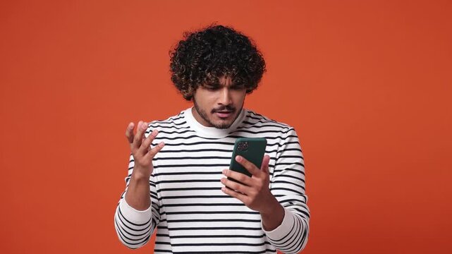 Sad upset young Indian man wear striped casual clothes hold use mobile cell phone swear read bad fake news unexpected rumor has some problem isolated on plain orange red background. Lifestyle concept