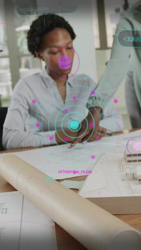 Vertical video: Seated designer studying plans, peer pointing, holo UI marking model, guiding edits