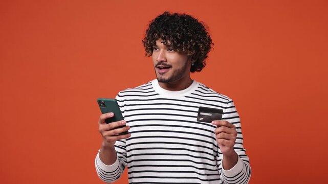 Young Indian man he wear striped sweatshirt casual clothes use mobile cell phone app hold in hand credit bank card doing online shopping order delivery to home isolated on plain orange red background