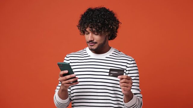 Young fun Indian man wear striped sweatshirt casual clothes use mobile cell phone app hold in hand credit bank card doing online shopping order delivery to home isolated on plain orange red background
