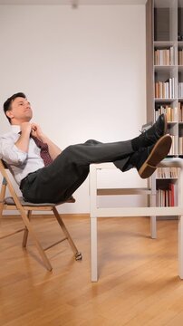 Vertical video businessman in business attire relaxing at his desk, having a break from work by leaning back in his chair with his feet on the table, reflecting or daydreaming
