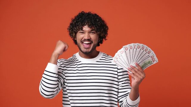 Young Indian man he wear striped sweatshirt casual clothes hold in hand fan of cash money in dollar banknotes doing winner gesture celebrate isolated on plain orange red background. Lifestyle concept