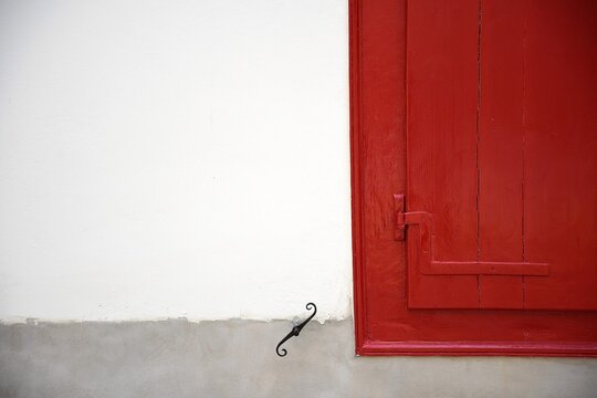 Traditional rural house window with crimson red wooden shutters and a forged iron shutter dog on a whitewashed wall in Galaxidi, Phocis Greece.