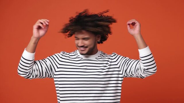 Young vivid fun Indian man he wear striped sweatshirt casual clothes dance waving rising have fun enjoy relax celebrate play fluttering hair isolated on plain orange red background. Lifestyle concept
