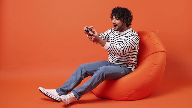 Young Indian man he wear striped sweatshirt casual clothes sit in bag chair hold in hand play pc game with joystick console do winner gesture isolated on plain orange red background. Lifestyle concept