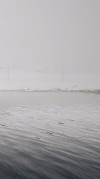 Fog covers the water and snow surrounds the area. The scene shows a blend of grayish tones as visibility is low. There is a calm water surface reflecting the foggy sky.