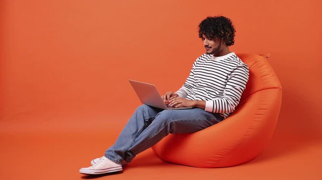Young programmer Indian IT man he wear striped sweatshirt casual clothes sit in bag chair hold use work on laptop pc computer isolated on plain orange red background studio portrait. Lifestyle concept