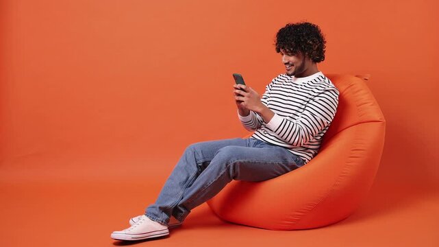 Young Indian man he wear striped sweatshirt casual clothes sit in bag chair hold in hand use point index finger on mobile cell phone isolated on plain orange red background studio. Lifestyle concept