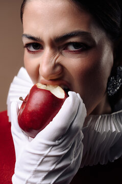 Portrait of a serious young woman in a red dress with a white collar and gloves eating a red apple