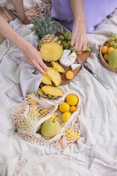 Overhead view of two people's hands reaching for slices of pineapple and camembert cheese on a picnic blanket