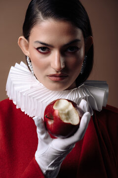 Portrait of a serious young woman in a red dress with a white collar and gloves holding a half eaten red apple