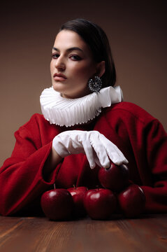 Close-up portrait of a serious young woman in a red dress with a white collar and gloves sitting at a table with a stack of red apples