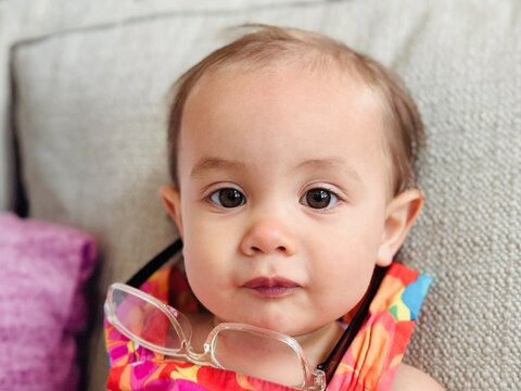 Portrait of a mischievous toddler sitting on a sofa with a pair of classes upside down around her neck