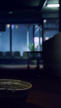 A metal bucket is placed on the floor in an indoor location with chairs in the background. Soft light enters through a window. The space appears empty and still at midday.