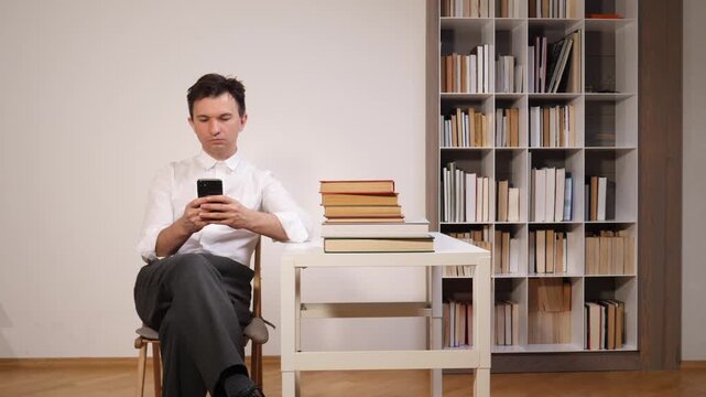 Man in white shirt seated at a wooden desk in a minimalist study, focused on his smartphone screen beside stacked books and a modern bookshelf, studying or researching indoors