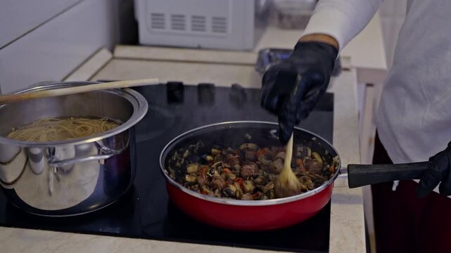 A person in black gloves skillfully stirs mushrooms, vegetables, and sausages in a red pan on a modern induction cooktop.