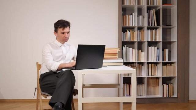 Man focusing on his laptop, working from a desk setup at home with a large bookshelf filled with books in the background, representing remote work and studying