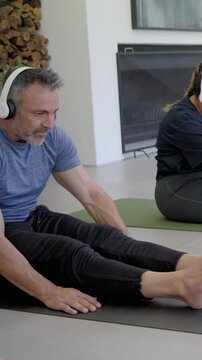 Vertical video: Leaning man in blue tee white headset reaching feet on black mat at home after cue