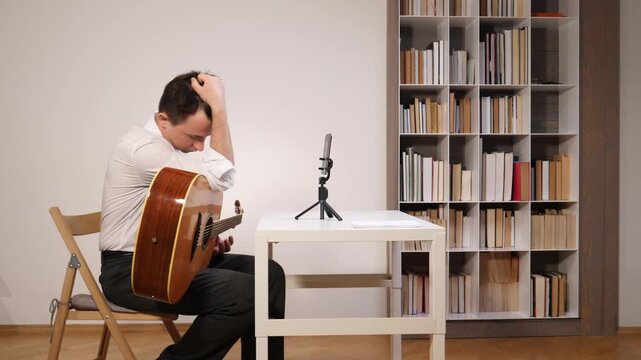 Man with an acoustic guitar sitting frustrated at a desk with a smartphone on a tripod, attempting to learn music through an online lesson in a home setting