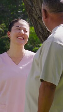 Vertical video: Smiling woman in pink scrub top telling joke, man in grey tee listening under tree