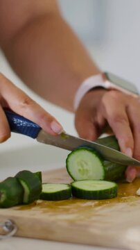 Vertical video: Hands slicing cucumber in kitchen left steadying, blue knife pink watch for salad