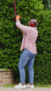 Vertical video: Raising red trimmer, man shaping hedge on walkway by planter, wearing gloves, muffs