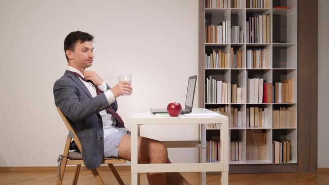 Man in a blazer, shirt, and tie without pants drinking water while using a laptop for a remote video conference, illustrating the new normal of working from home with a humorous twist
