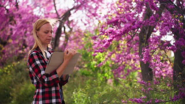 Botanist researching flowering trees using tablet in natural environment. Scientific study of plants, botany concept, spring blossom, field research and data collection outdoors.