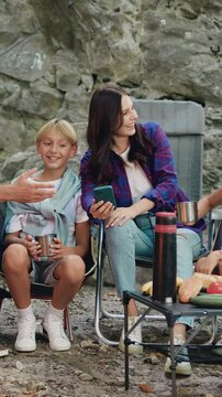 Happy young handsome parents and their children using smartphone watching video or browsing internet together online sitting near campfire during weekend outdoors in nature. Leisure and technology