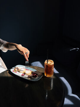 Side view of a woman sitting at a table eating mini Syrniki pancakes with yogurt and blueberries served with a refreshing iced espresso tonic
