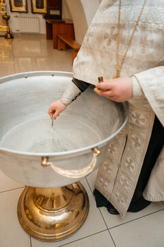 An Orthodox priest performing the ritual of blessing the water in a large silver baptismal font. He uses a special tool to drop holy oil into the water. Symbolic sacrament of baptism 