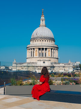 View of a woman in a flowing crimson dress admiring the majestic, weathered dome of St. Paul's Cathedral under a brilliant blue sky, London, England, United Kingdom.