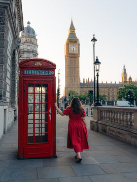View of a woman in a scarlet dress pausing by a vivid red telephone box, with the timeless Big Ben rising majestically under a soft sky, London, England, United Kingdom.
