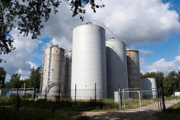 Fototapeta premium Industrial storage tanks under a cloudy sky, surrounded by trees and fencing