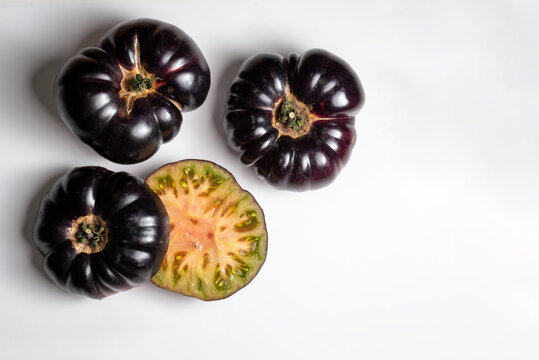 Toma a&eacute;rea de tomates negros Indigo Rose, incluyendo un corte transversal que muestra su estructura interna. Composici&oacute;n minimalista con espacio negativo sobre una superficie blanca con espacio para t