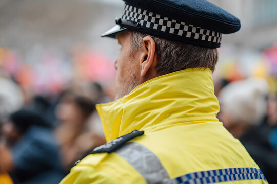 Yellow jacket, checkered cap. Back view, focused on shoulder gear. Crowd blurred, suggests public event. Neutral expression, duty-focused gaze. Ideal for safety, authority, civic scene