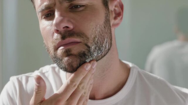 Facing bathroom mirror man checking and rubbing jaw with right hand, assessing stubble in white tee