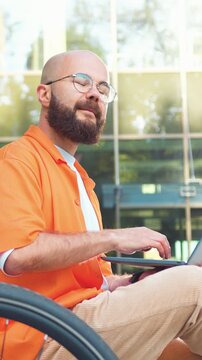 Cheerful smiling bearded man in glasses typing on keyboard sitting on bench with his bicycle beside him outdoor. Handsome guy working on laptop computer near modern glass office. Slow motion.