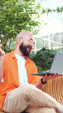Stylish hipster man in glasses working in laptop while sitting in city square on bench with bicycle at sunset. Freelancing and outdoor education concept. Slow motion.