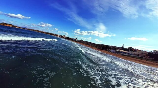 Aerial FPV view of the vibrant blue sea crashing onto the warm reddish-brown sandy beach, with lush green palm trees under a bright sky, Galatas, Chania, Greece.