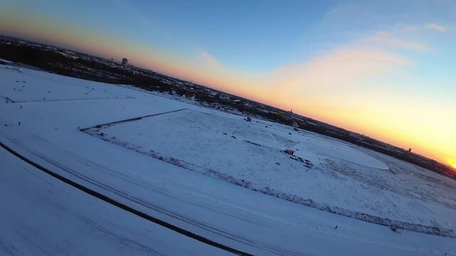 Aerial FPV view of a vast, pristine white snow-covered field with distant buildings and trees under a deep blue sky, Berlin, Berlin, Germany.