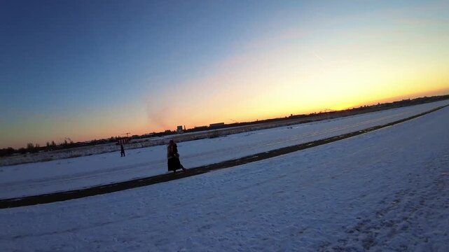Aerial FPV view of a solitary figure traversing the vast, snow-dusted expanse of Tempelhofer Feld under a vivid, gradient sky, Berlin, Berlin, Germany.