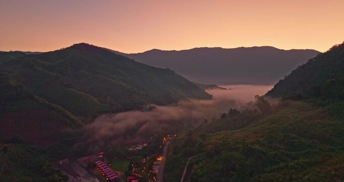 A serene aerial view of a mountain valley at sunrise, with soft mist drifting through the hills and warm lights glowing from a small village below. The scene captures the peaceful atmosphere