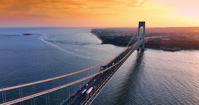 Beautiful scenery of the Verrazzano-Narrows Bridge with lively traffic. Stunning orange sky over the waterscape and cityscape in the backdrop. Aerial view.