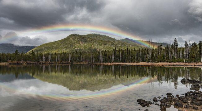 Rainbow in dark clouds over a forest, reflected in Sparks Lake, Oregon, USA