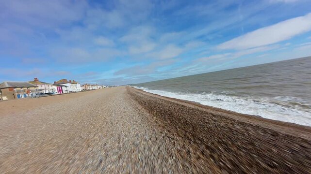 Aerial FPV view of the expansive, textured pebble beach meeting the foaming, grey-blue sea under a bright, cloudy sky, with a vibrant row of houses, Aldeburgh, England, United Kingdom.